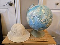 Photo of blue vintage globe on olive green metal stand and white mesh safari hat both on wooden surface near door handle.