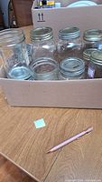 Photo showing multiple clear glass mason jars in a cardboard box, different sizes and some with metal lids removed or not visible