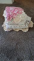 Stack of assorted crochet and lace doilies on carpet showing varied sizes and colors including white, cream, and pink