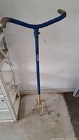 Full view of the blue and yellow metal garden claw tool standing upright, showing dual handles and metal claw at bottom.