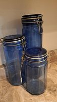 Photo showing three blue glass canisters of varying heights with octagonal shapes and metal clamp lids on a countertop.