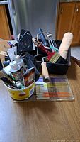 Overview of painting tools set including paintbrushes, foam roller, masking tape, small paint containers, and paint tray on wooden table.