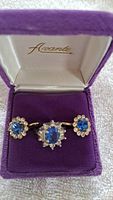 Jewelry set comprising one silver ring with oval sapphire and two matching silver earrings with round sapphire stones, displayed in a purple velvet Avante-branded box.