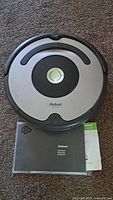Top view of iRobot Roomba robotic vacuum cleaner placed on carpet with user manuals underneath.