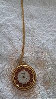 Front view of Lucerne vintage necklace watch pendant showing white dial, red numbered circular hour markers from 1 to 12, gold-tone rhodium plated casing, and chain lying on fabric background.