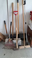 Photo showing garden rake, leaf rake, shovels, hoe, and sledgehammer with wood handles and metal parts, arranged against wall