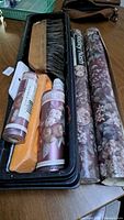 Photo of two rolls of floral patterned Ridley Nash vinyl wallpaper, and wallpaper tools including a brush and smoothing tool in a black plastic tray on a wooden table.