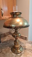 Photo showing vintage brass electric table lamp with dome-shaped brass shade and small glass chimney on top. Base is detailed with decorative rounded brass elements. Lamp cord visible.