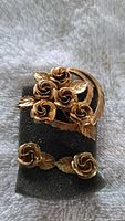 Gold-tone metal vintage rose flower brooch with five roses and leaves, along with matching single rose flower clip-on earrings each with a leaf, displayed on black foam.