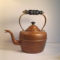 Front view of the antique copper tea kettle showing its aged surface, black painted turned wooden handle, and spout with hinged lid.
