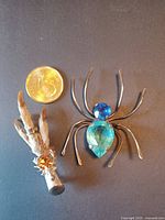 The two pins shown together with a coin for scale: a silver spider brooch with blue stones and an antique grouse foot pin with an orange stone.