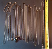 Sixteen delicate chains and bracelets arranged against black background with ruler for scale