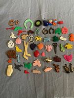 Full set of 53 colorful small plastic prizes and charms spread on a gray fabric background, including animals, horseshoes, boots, sports items, fruits and others.