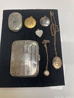 All items arranged on black mat showing two silver cases at left and five lockets at right