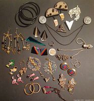 Photo showing 30+ assorted costume jewelry pieces arranged on a dark surface showing a wide variety of items including earrings, pins, bangles, geometric shapes, animal motifs and ornate designs