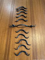 Eleven hand-forged cabinet handles arranged on a wooden surface, ten smaller curved handles and one longer handle with curled ends.