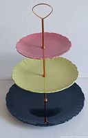 Assembled 3-tier serving tower showing scalloped edge pink (top), yellow (middle), and navy blue (bottom) plates with copper-tone metal support and handle.