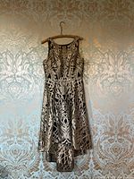 Full front view of sleeveless black dress with gold embroidery hanging on a wooden hanger against a patterned beige and silver wallpaper.