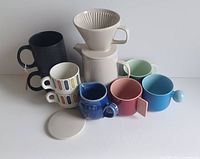 Front view photo showing all items in the set including coffee pot, filter cone, lid, seven mugs in various colors and styles, and a cream pitcher.