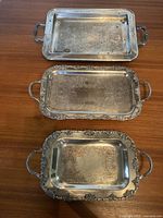 Photo showing three silver trays arranged vertically, highlighting etched details and ornate grape-motif edges on two trays.