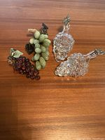 Photo showing two clear glass grape-shaped bottles with cork and metal leaf stoppers alongside red and green glass grape bunches with carved leaves.