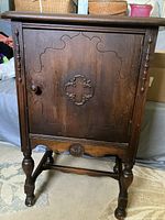 Front view of vintage solid wood cabinet showing ornate carved single door with round wooden knob and detailed legs.
