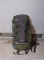 Front view of the green and grey Mountain Equipment Co-op IBEX 65L hiking backpack on a blank indoor floor against a wall.