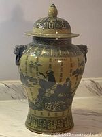Front view of antique oriental porcelain urn with lid showing hand painted blue figures and calligraphy on beige background with elephant handles.