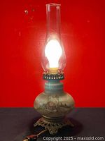 Front view of antique kerosene table lamp with clear glass chimney, showing hand-painted countryside scene on milk glass body and ornate brass base with power cord.