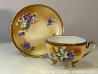 Front angle of teacup showing hand painted floral design, three feet, and saucer with matching gold rim