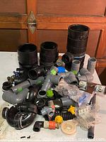 Top view of the assorted plumbing parts on a white surface including black pipe couplings, gray PVC elbows, valves, and other connectors.