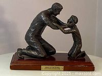 Full view of bronze sculpture showing father and son kneeling, mounted on a wooden base with plaque.