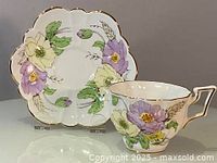 Hand-painted teacup and matching saucer with large purple and white floral motifs, vivid green leaves, scalloped edges, and gold trim