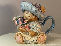 Front view of vintage Fitz and Floyd bear teapot holding flowers in detailed blue hat and decorative clothing.