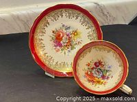 Teacup and saucer set showing multicolored hand painted flowers, gold filigree decoration, and deep red border on white china.
