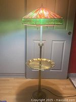 Full view of vintage slag glass floor lamp lit, showing solid brass base and attached brass tray, standing on floor in front of closed door.