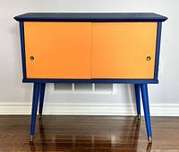 Front view of vintage mid century modern wooden record cabinet painted cobalt blue with orange sliding doors on hardwood floor.