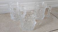 Three clear glass mugs showing embossed 3D character designs of Batman, Robin and Riddler from the 1995 McDonald's Batman Forever promotion lying on tile surface.
