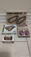 Photo of the original sealed Tamiya Track & Wheel Set box and boxes of the motor, gearbox, and sports tire set placed on floor against wall.