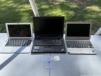All three laptops shown open on a white table outdoors: Apple MacBook Air (left), Lenovo ThinkPad (center), and Samsung (right).