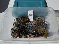 Overhead view of clear plastic bin filled with assorted jewelry items including multiple bracelets, necklaces, earrings, and brooches in various colors and styles