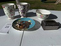 Two floral ceramic cups, small floral bowl, metal trinket box, and decorative coin dish with coins displayed on a white table outside