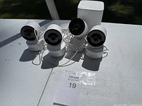 Four white wired home security cameras and one white base station placed on a white outdoor surface in daylight.