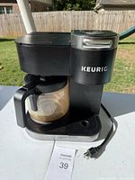 Front and side view of black Keurig coffee maker with glass carafe on a white table outside.