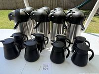 Set of 10 stainless steel airpot coffee dispensers with black handle and top, arranged in two rows outdoors on grass.