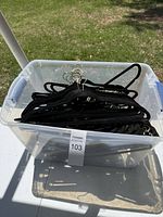 Clear plastic bin filled with black velvet felt hangers with silver metal hooks inside, photographed outdoors on grass.