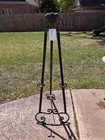 Front view of the tall metal art easel standing upright on concrete, showing detailed scrollwork decoration and overall structure.