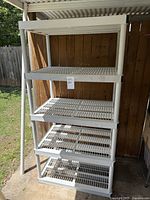 Front and side view of a large white plastic shelving unit with five open grid shelves placed outdoors by a wooden fence and covered patio area.