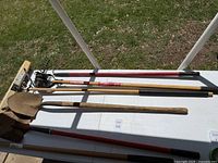 Top view of yard tools laid out on a white surface showing two shovels, a rake, a hoe, and additional hand tools including a Craftsman branded tool.