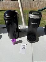 Front view of a black Nespresso coffee machine, a clear glass, a black Keurig coffee machine, and a used purple coffee capsule on a white table outdoors.